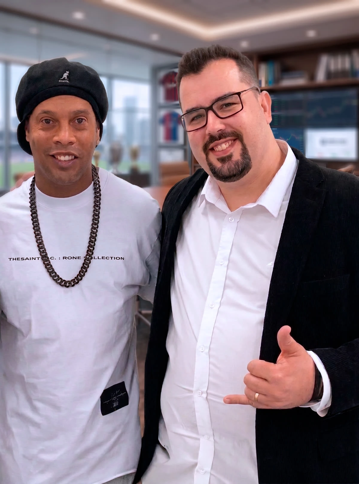 Lucas Correa ao lado de Ronaldinho Gaúcho, em escritório com camisas de futebol emolduradas e telas de mercado financeiro ao fundo.
